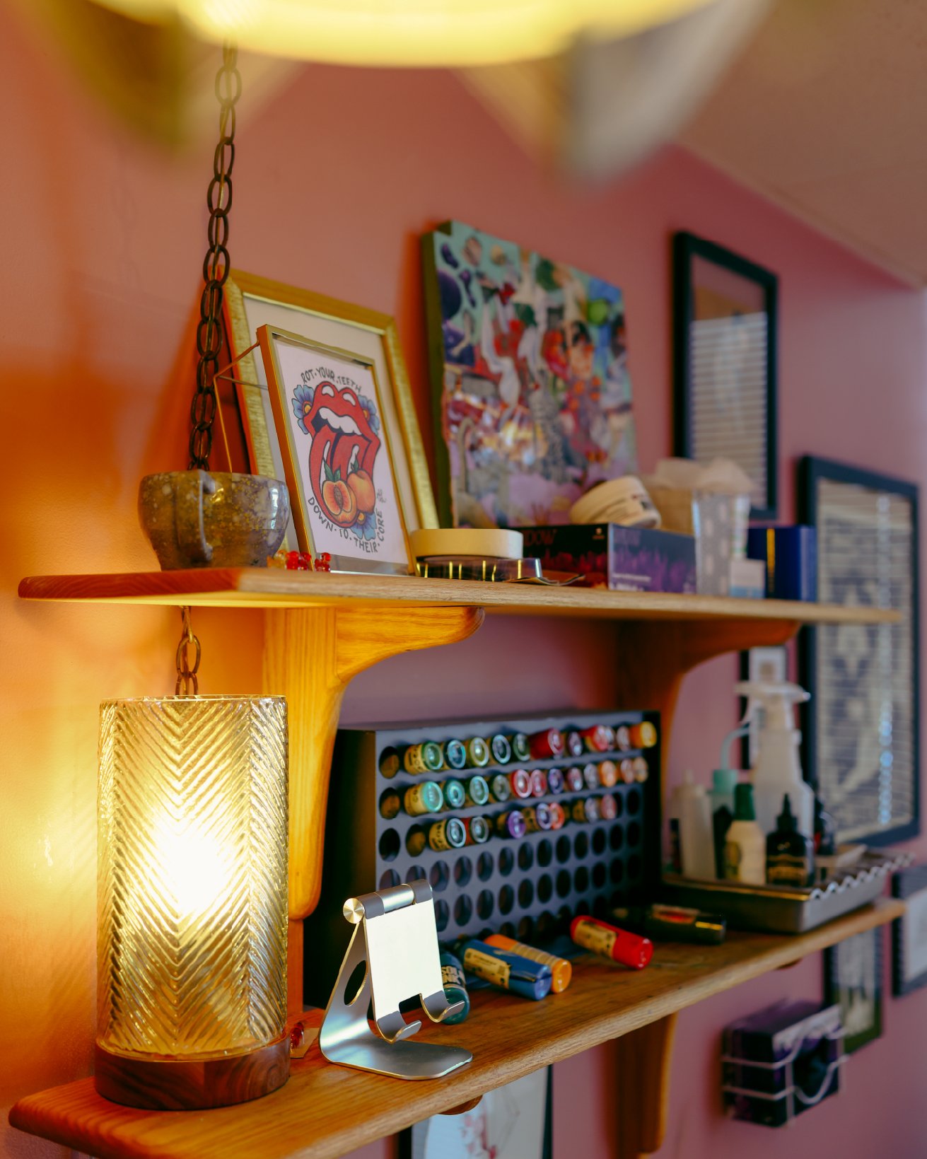 A wooden shelf with a decorative lamp, framed artwork, and various art supplies, with a pink wall and mirror in the background.