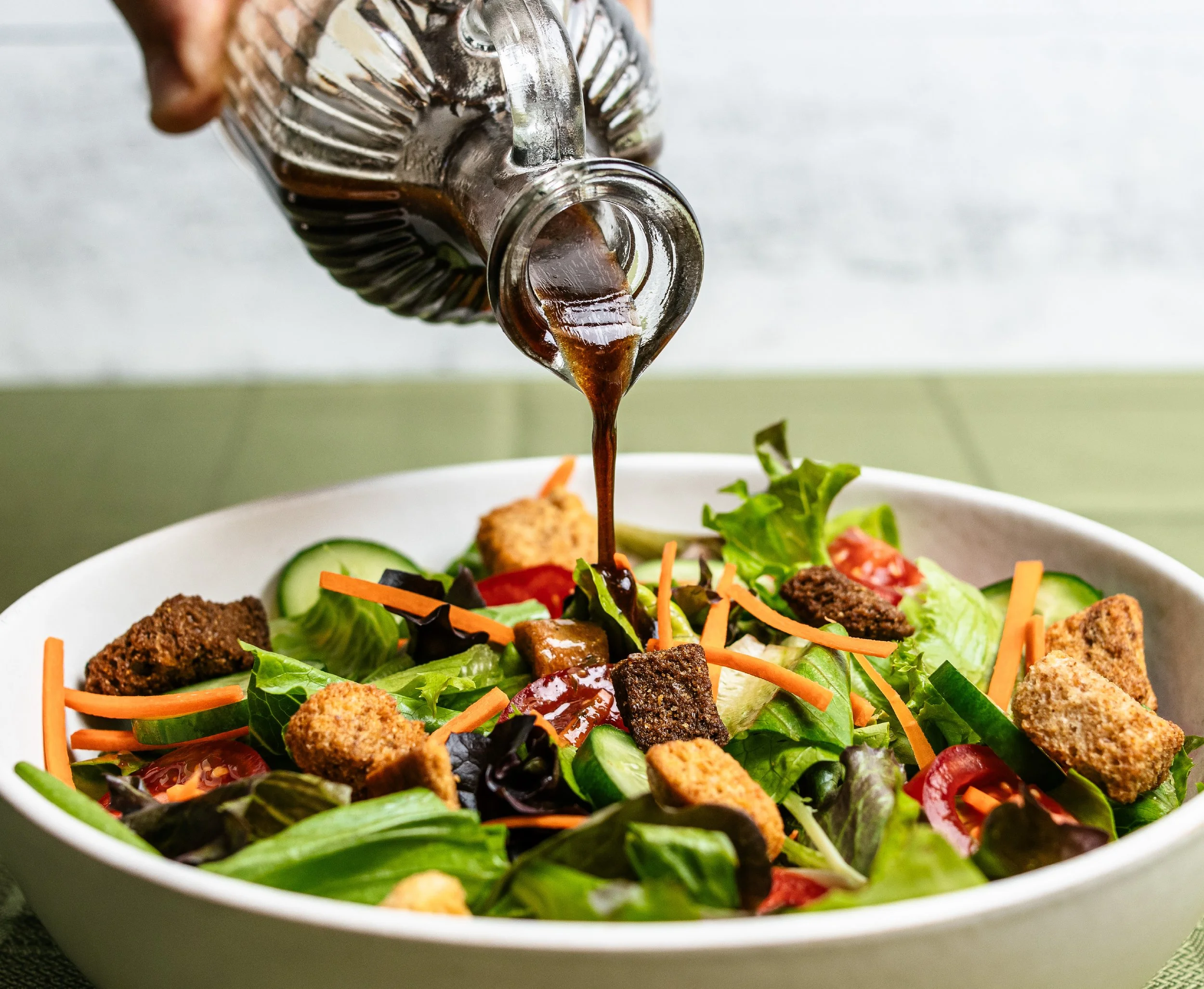 A salad with lettuce, cherry tomatoes, shredded carrots, cucumber slices, croutons, and crispy bacon bits. A sauce or dressing is being drizzled onto the salad from a glass bottle.