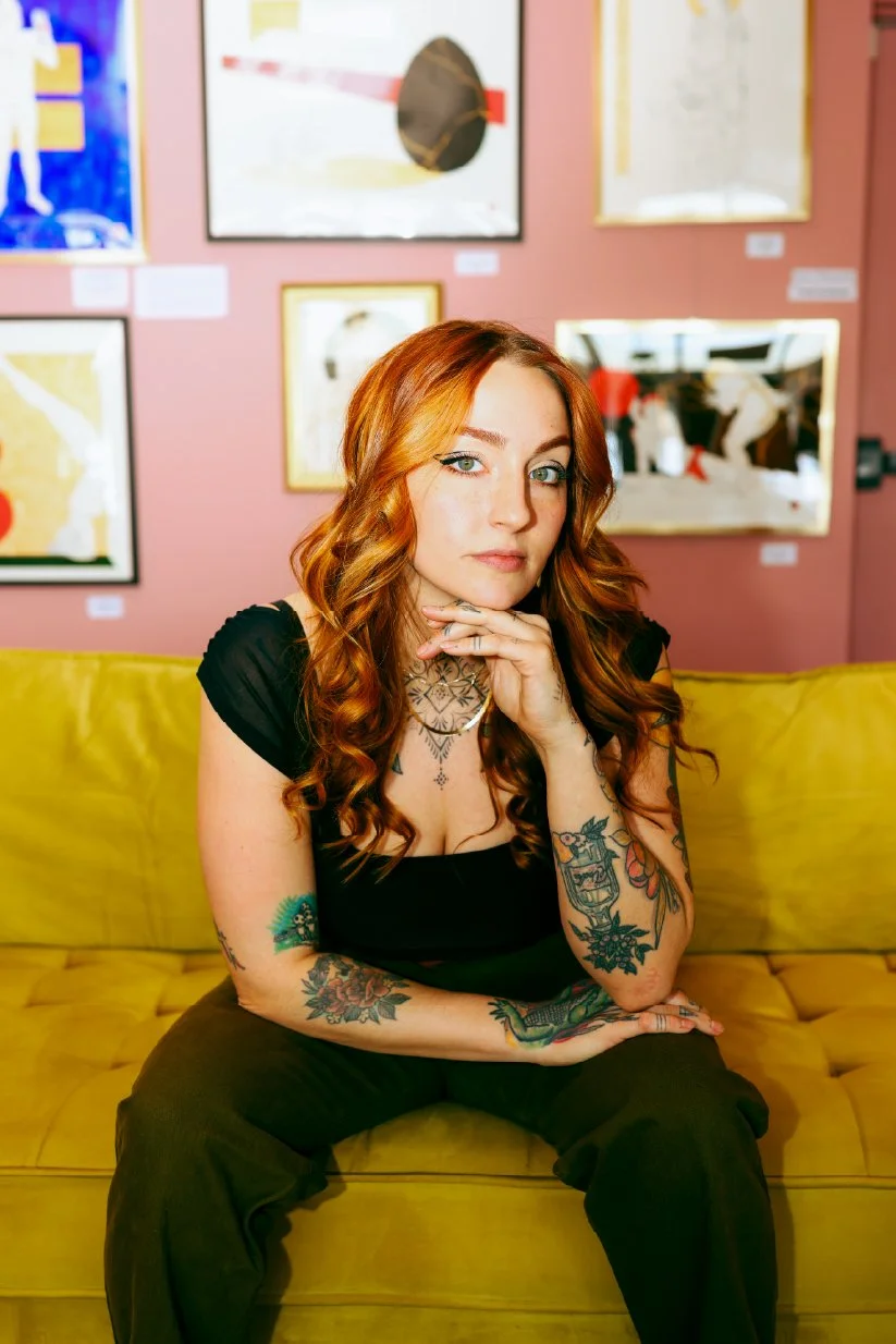 A red-haired woman with tattoos, wearing a black top, sitting on a yellow couch in an art gallery with various framed artworks on a pink wall behind her.