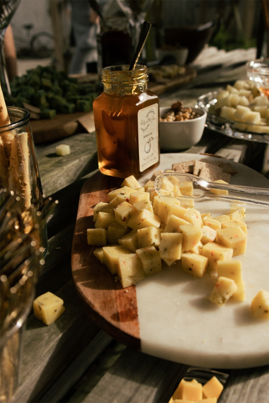 Cheese cubes on a marble platter with a jar of honey, a cheese knife, and various cheeses and snacks on a wooden table in an outdoor setting.