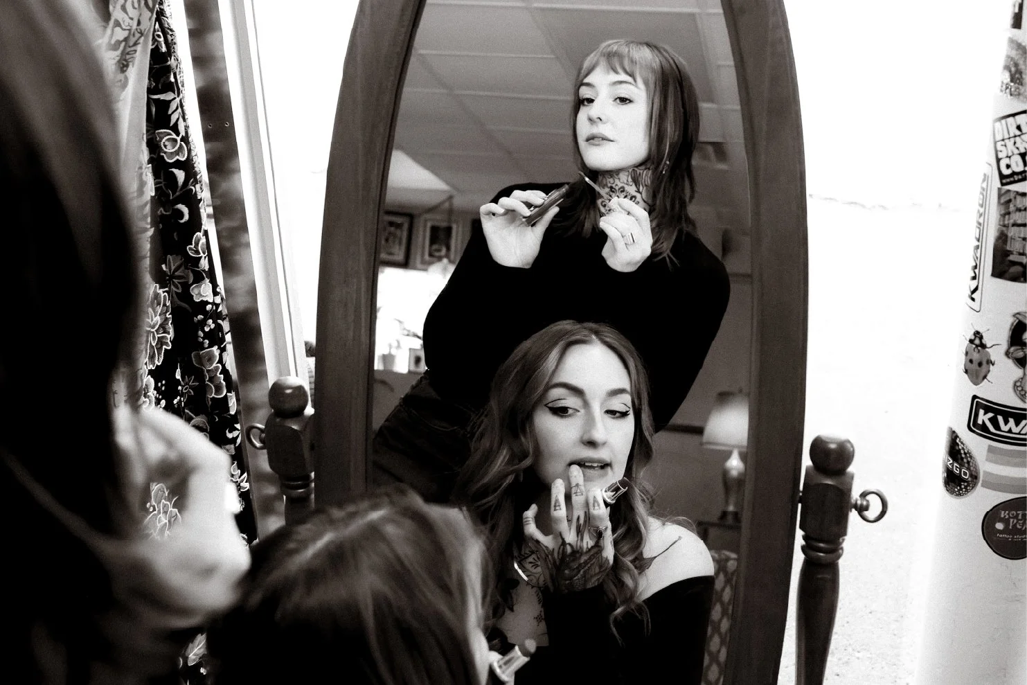 A young woman with tattoos on her hand is applying makeup while looking in a mirror, with her reflection also shown in the mirror. She has long hair and is sitting at a vanity table.