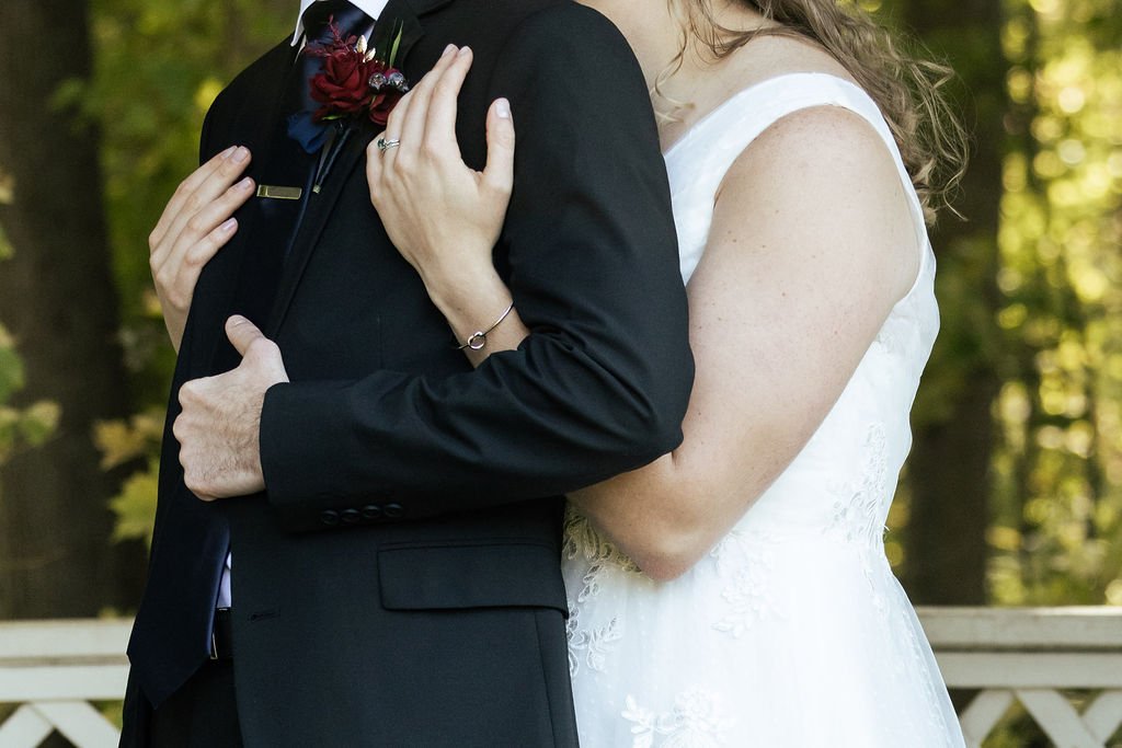 Close-up of a bride and groom embracing outdoors, with only their torsos visible. The groom is wearing a black suit with a red boutonniere, and the bride is in a white wedding dress with lace details.