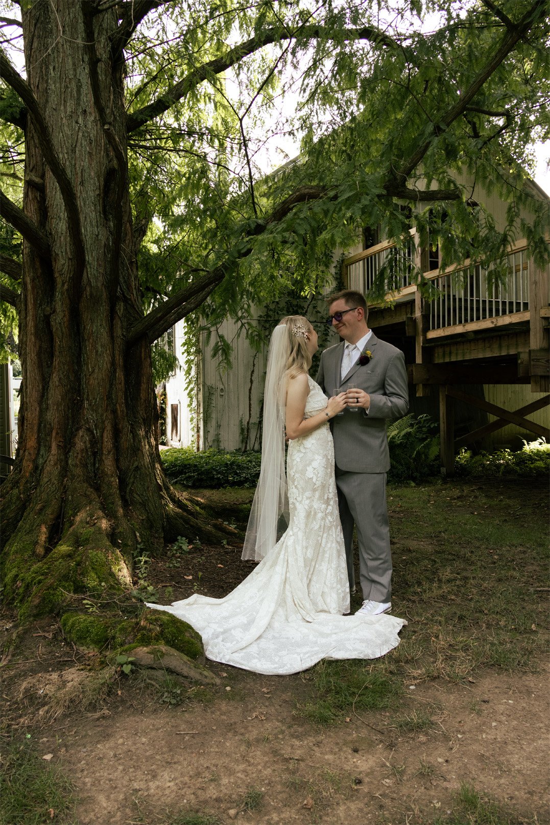 Bride and groom standing close under a large tree, holding hands, with the bride wearing a white lace wedding gown with a long train and veil, and the groom in a gray suit, outside during daytime.