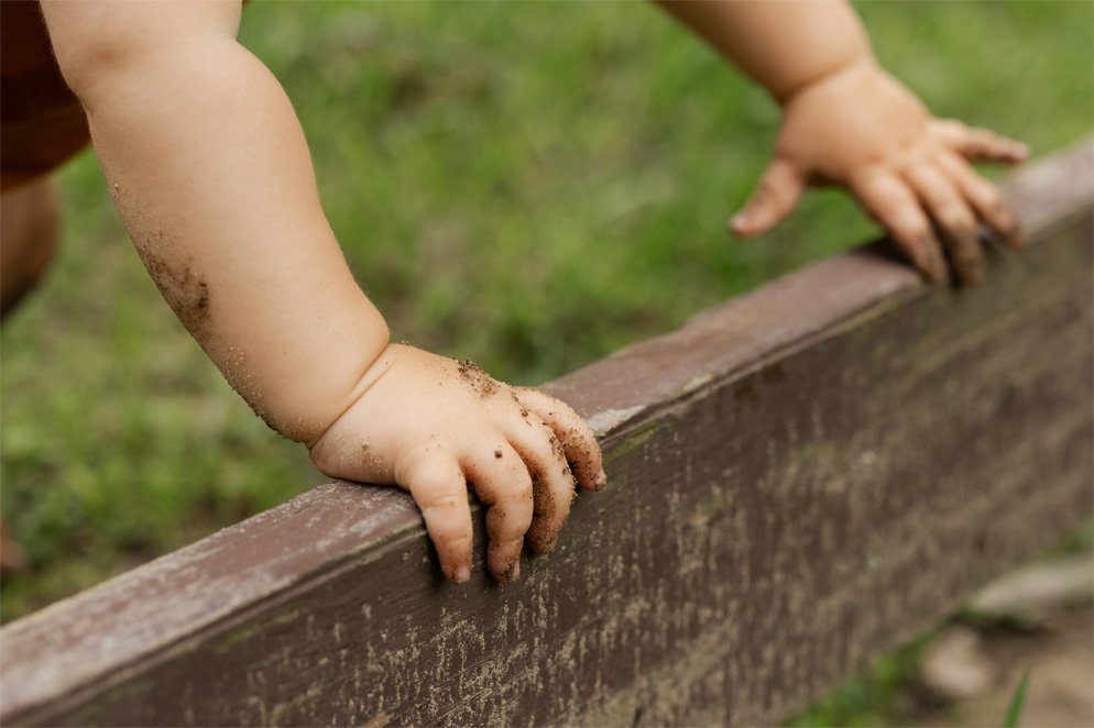 Close-up of a child's hands gripping a wooden fence, with dirt on their hands and arms, outdoors on grass.