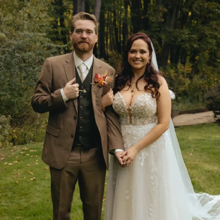 A groom and bride standing together outdoors in a forested area. The groom is dressed in a brown suit with a white shirt and tie, while the bride wears a white lace wedding dress and has long, dark hair. They are holding hands and smiling at the camera.