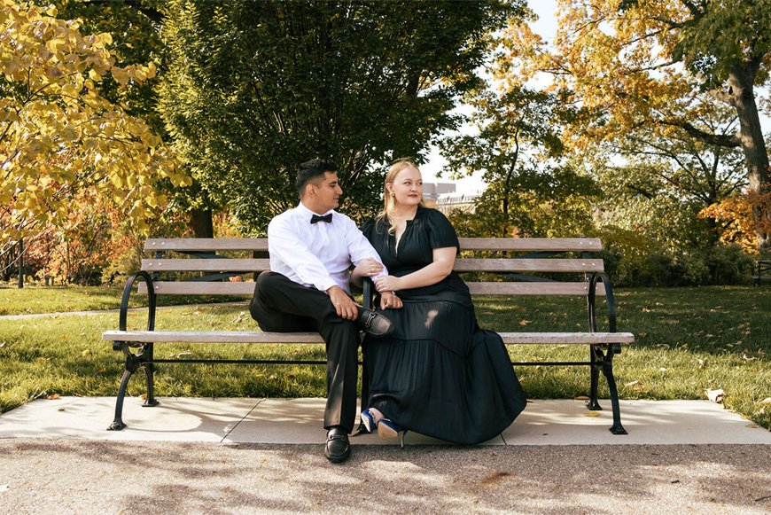 A man and woman sit on a park bench during autumn, holding hands and looking peacefully into the distance. The man wears a white shirt with a black bow tie, and the woman wears a long black dress. There are trees with changing leaves in the backgroun
