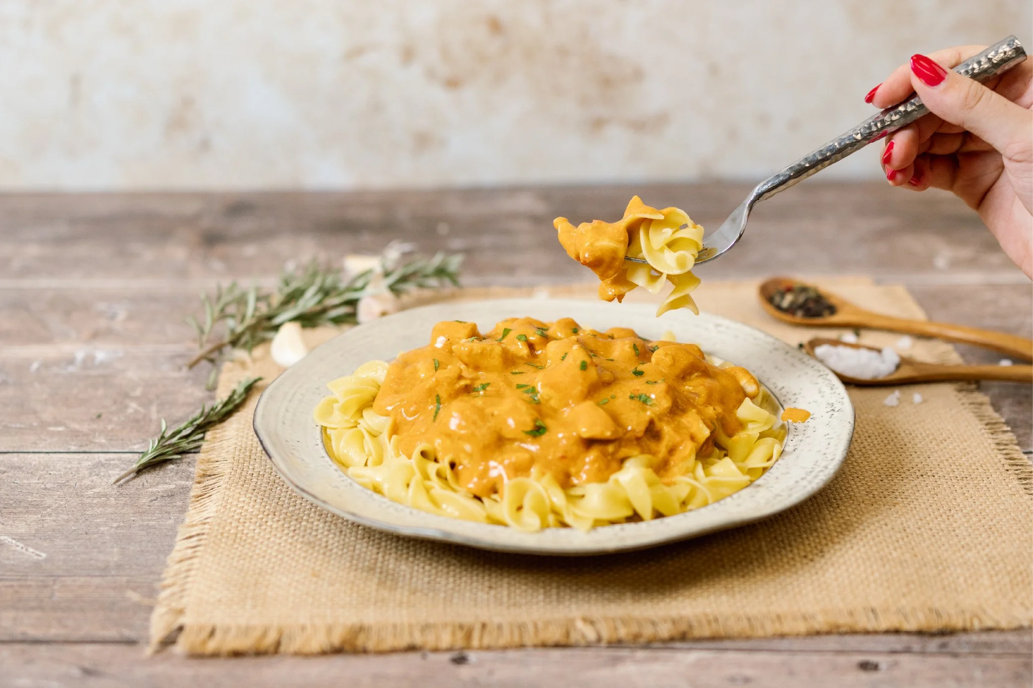 A plate of pasta topped with a creamy sauce with chunks of meat, garnished with chopped herbs, on a rustic wooden table with sprigs of herbs and garlic nearby.