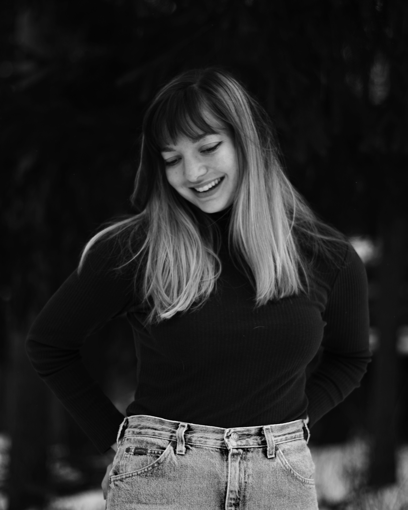 Black and white photo of a young woman with long hair, smiling, wearing a black top and high-waisted jeans, standing outdoors.