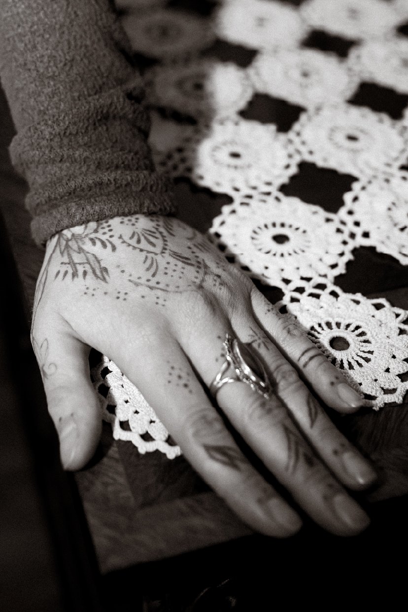 Close-up of a person's hand with tattoos, rings, and a long-sleeve shirt, resting on a lace doily on a wooden surface.