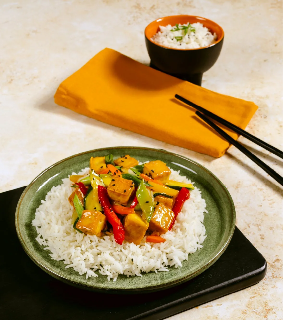 A plate of stir-fried vegetables with tofu over white rice, with a bowl of rice, chopsticks, and a folded orange napkin in the background.