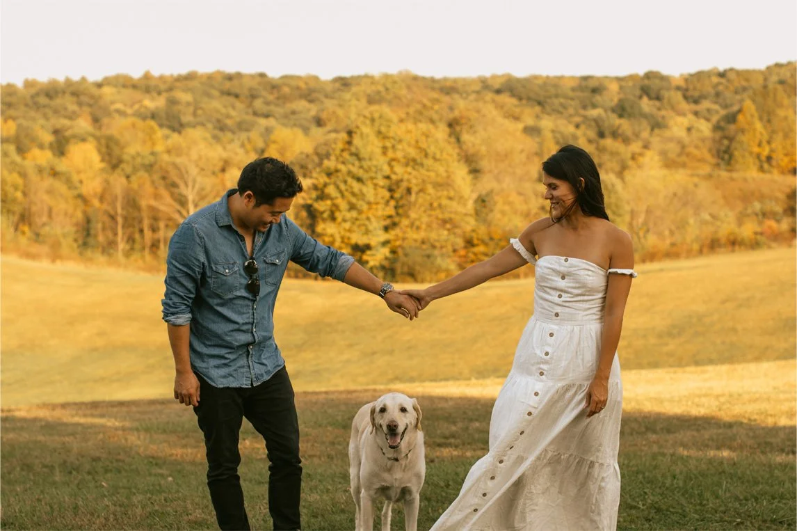 A couple holding hands with a Labrador Retriever dog on a grassy field with trees in autumn colors in the background.