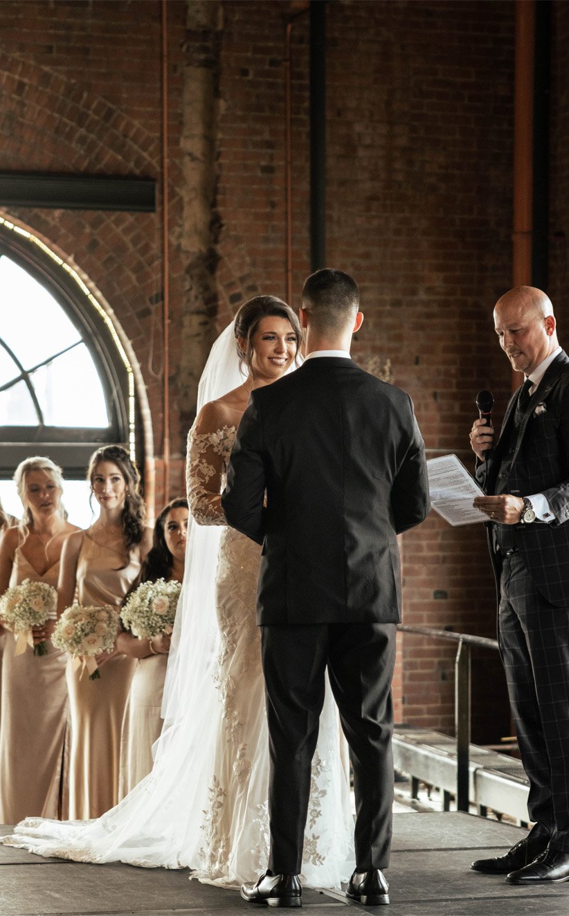 Bride and groom exchanging vows during their wedding ceremony inside a brick building, with bridesmaids holding bouquets in the background.