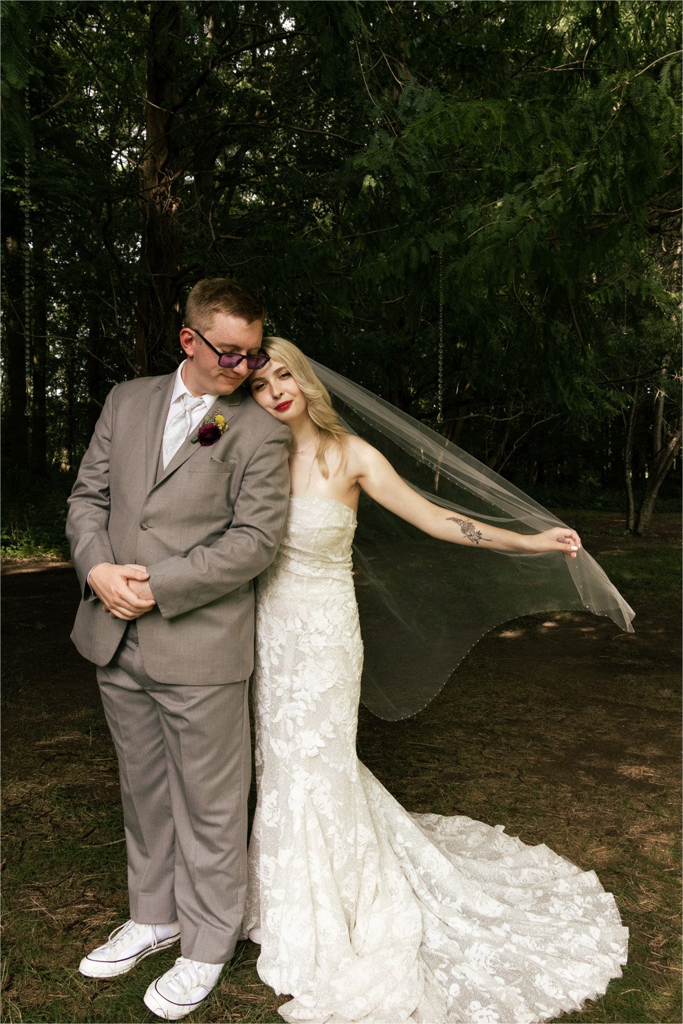 A bride and groom standing close together outdoors among trees, with the bride resting her head on the groom's shoulder. The bride is wearing a strapless lace wedding gown with a veil, and she has blonde hair and red lipstick. The groom is wearing a