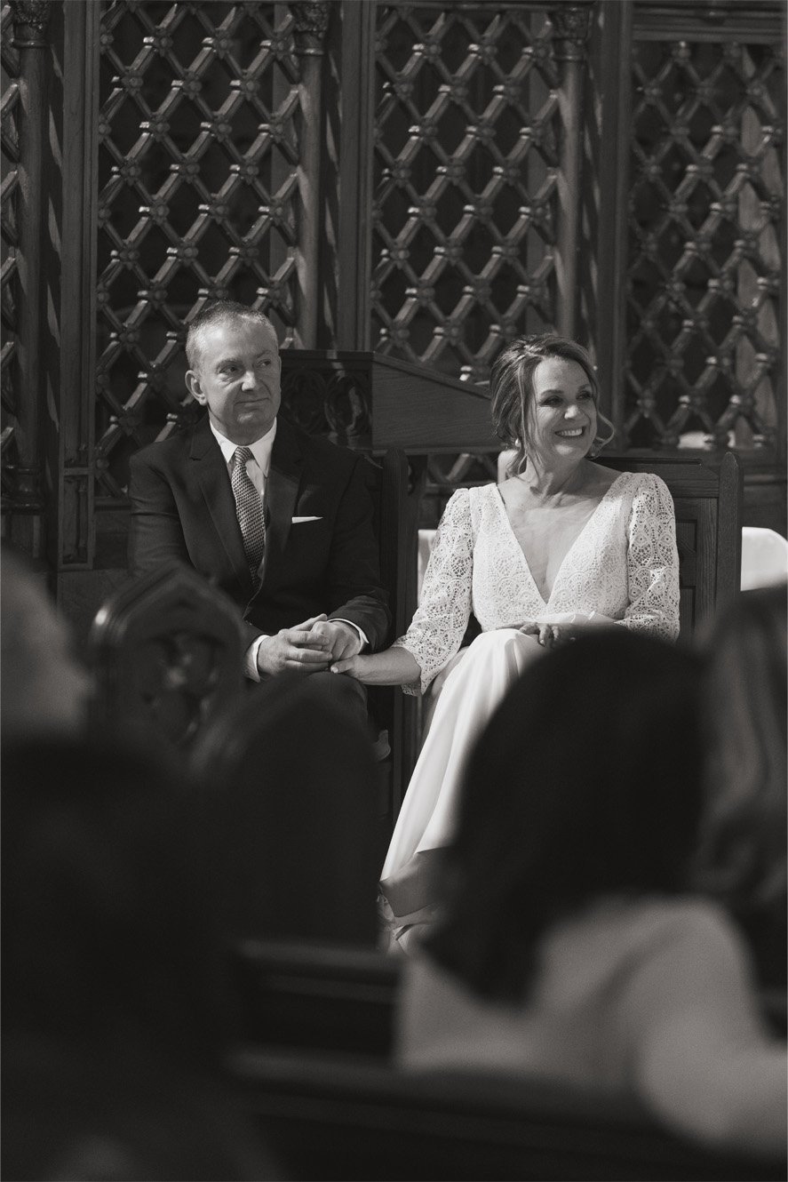 Black and white photo of a couple sitting in a church during a wedding ceremony, holding hands. The woman has shoulder-length hair and is wearing a lacy dress, smiling. The man has short hair and is wearing a suit, looking serious. The background fea