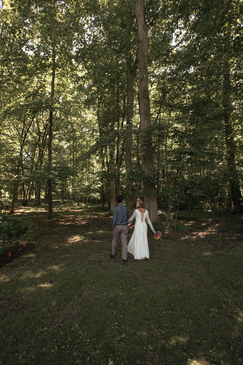 A bride and groom walk hand in hand through a forest with tall trees and dappled sunlight, the bride holding a bouquet of colorful flowers.