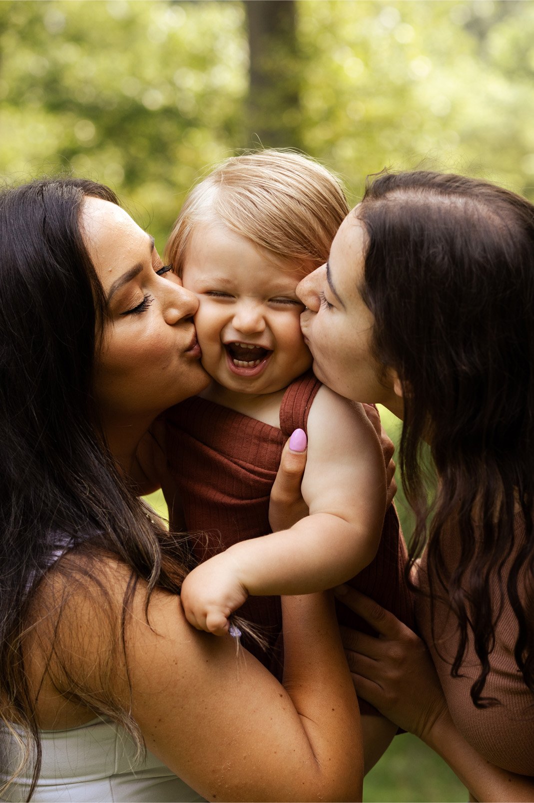 Two women with dark hair kiss a young girl with light brown hair on each cheek as she laughs outdoors in a green forest setting.