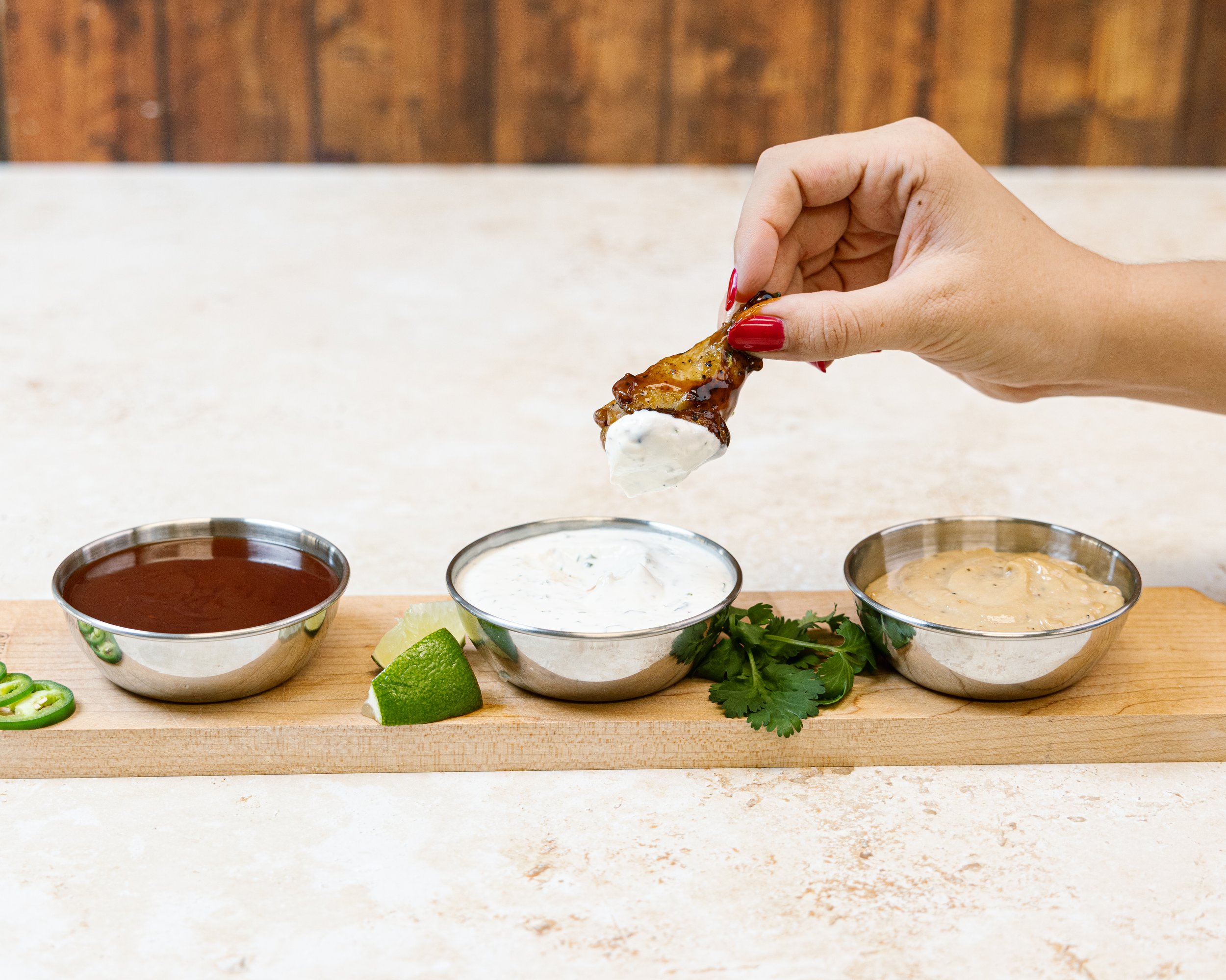A hand with red nails holding a piece of grilled chicken wing near three bowls of dipping sauces on a wooden surface, with lime wedges and cilantro leaves nearby, against a wooden background.
