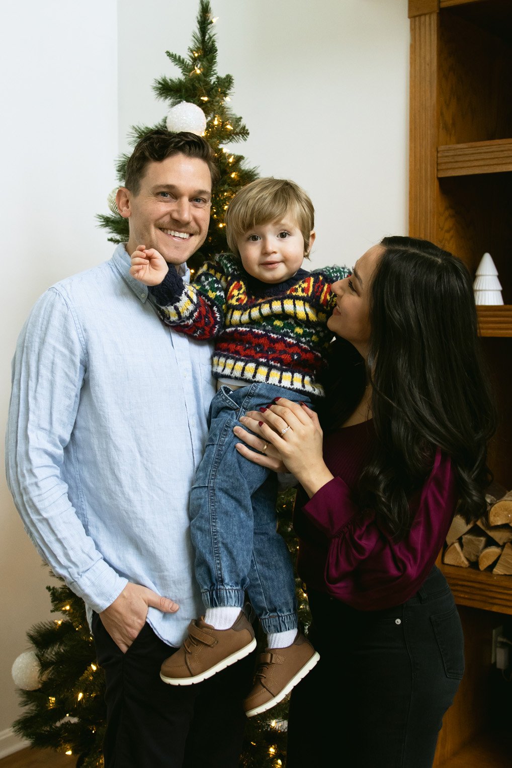 A family of three celebrating Christmas indoors, with a decorated Christmas tree in the background. The father, mother, and young son are smiling and enjoying the moment.