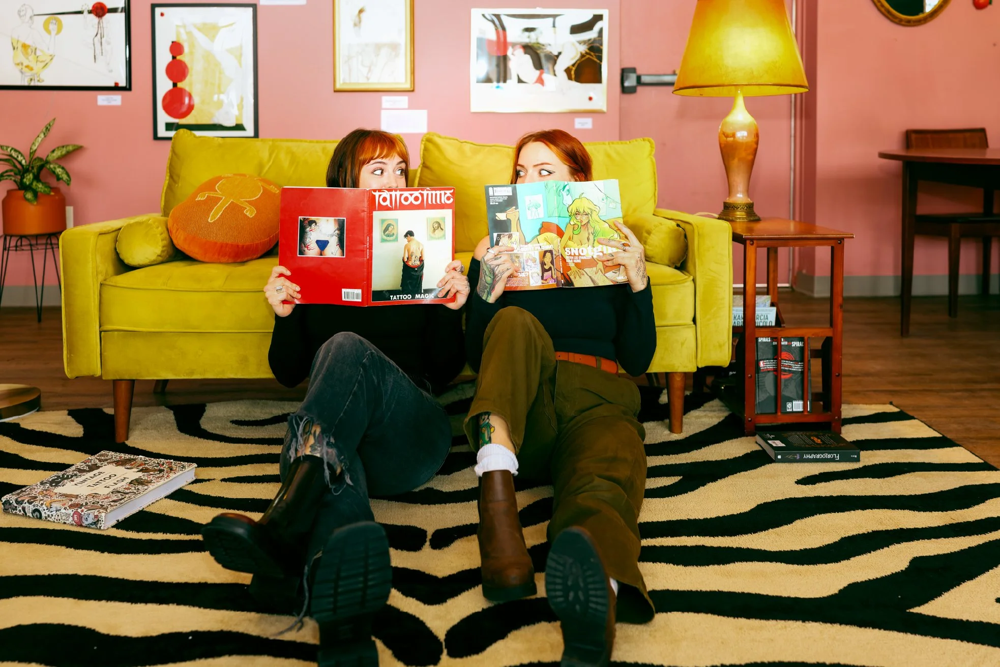 Two women with red hair sitting on a yellow couch in an art gallery, holding comic books and looking at each other, with a black and white striped rug and various framed artworks on the pink wall behind them.