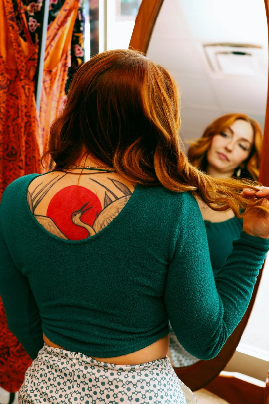 A woman with red hair looking into a mirror, adjusting her hair, revealing a back tattoo of a crane with a red sun background.