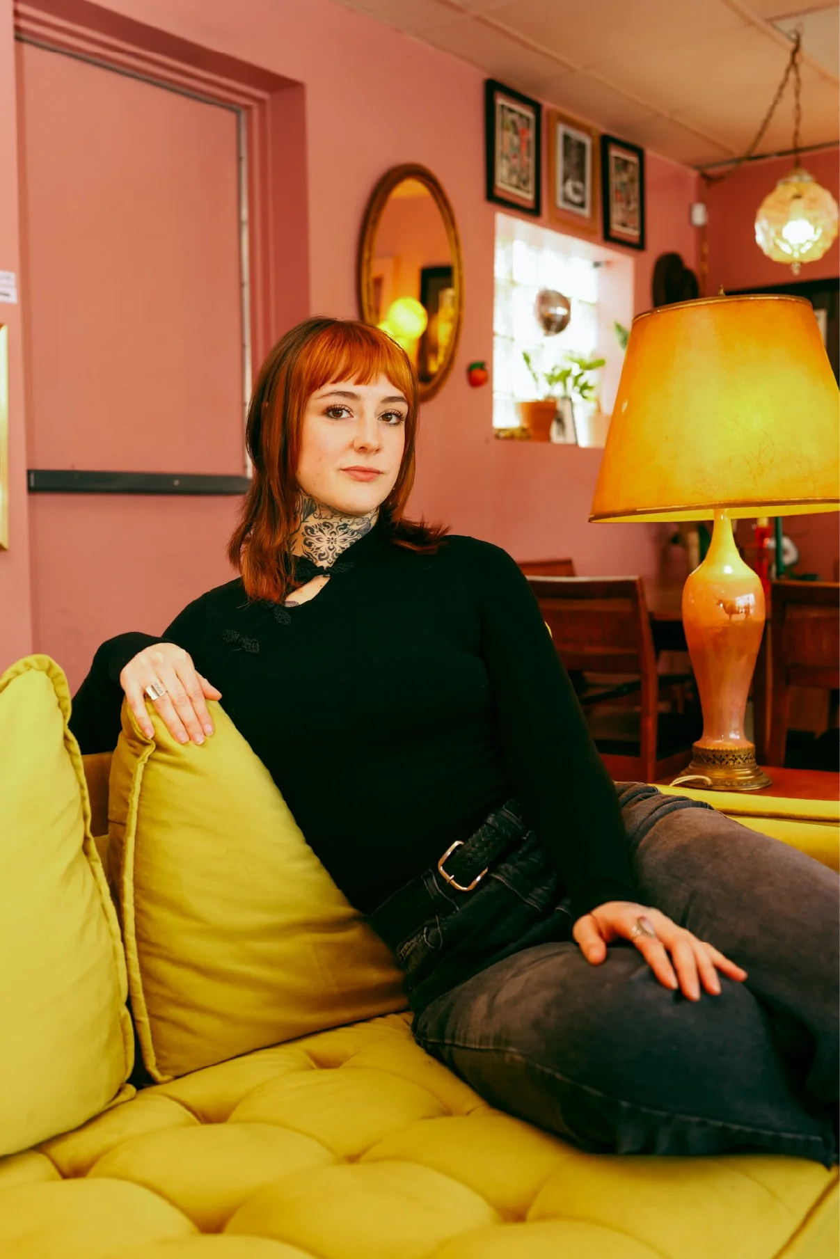 A woman with red hair and tattoos on her neck sitting on a yellow couch in a colorful, cozy room, with pink walls, framed pictures, a mirror, a window with plants, a yellow lamp, and wooden furniture.