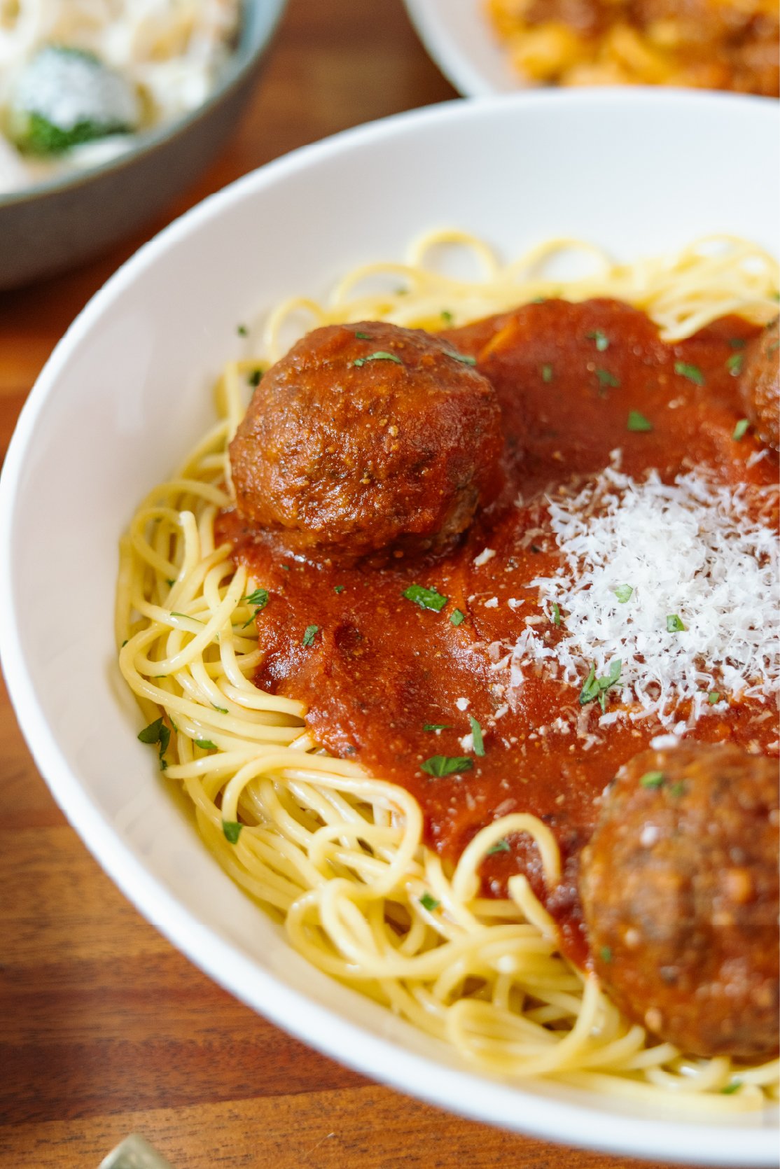A close-up of a bowl of spaghetti topped with meatballs, tomato sauce, grated cheese, and chopped herbs.