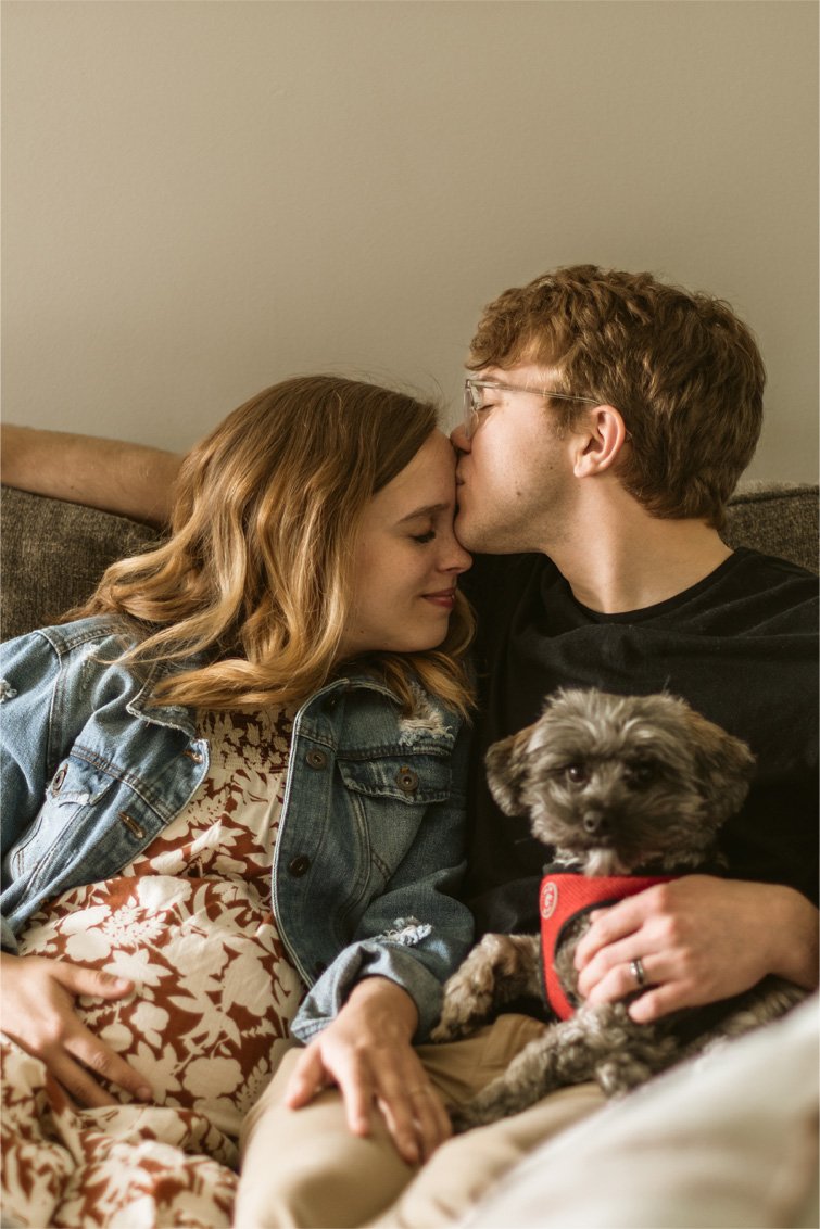 A couple sitting on a couch, with the man gently kissing the woman's forehead, while holding a small dog in his lap.