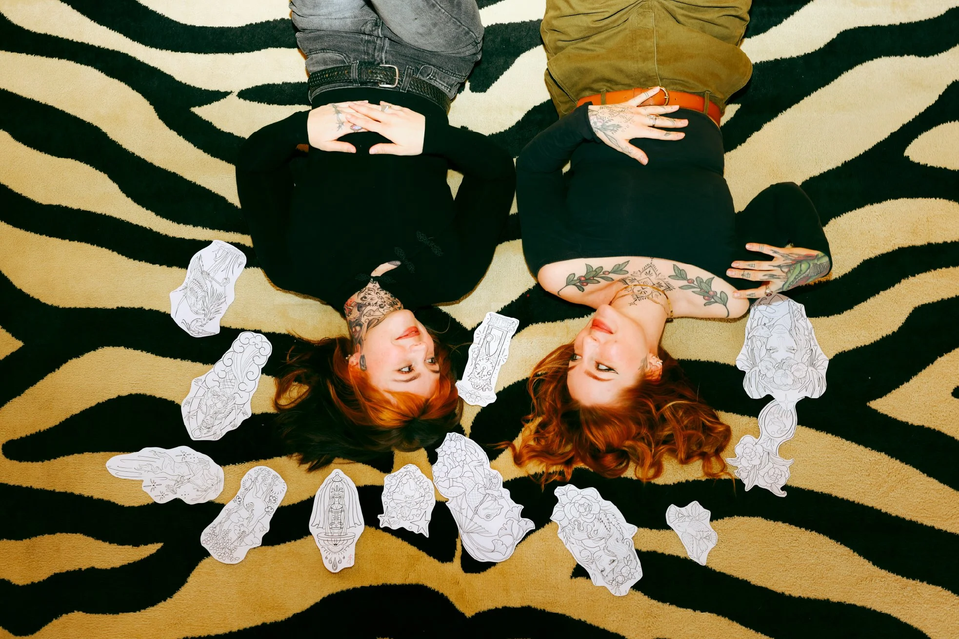 Two women lying on a black and beige zebra-striped rug with various black-and-white sketches of faces, hands, and other art pieces around their heads. One woman has dark hair, tattoos, and a black top, while the other has red hair, tattoos, and a bla