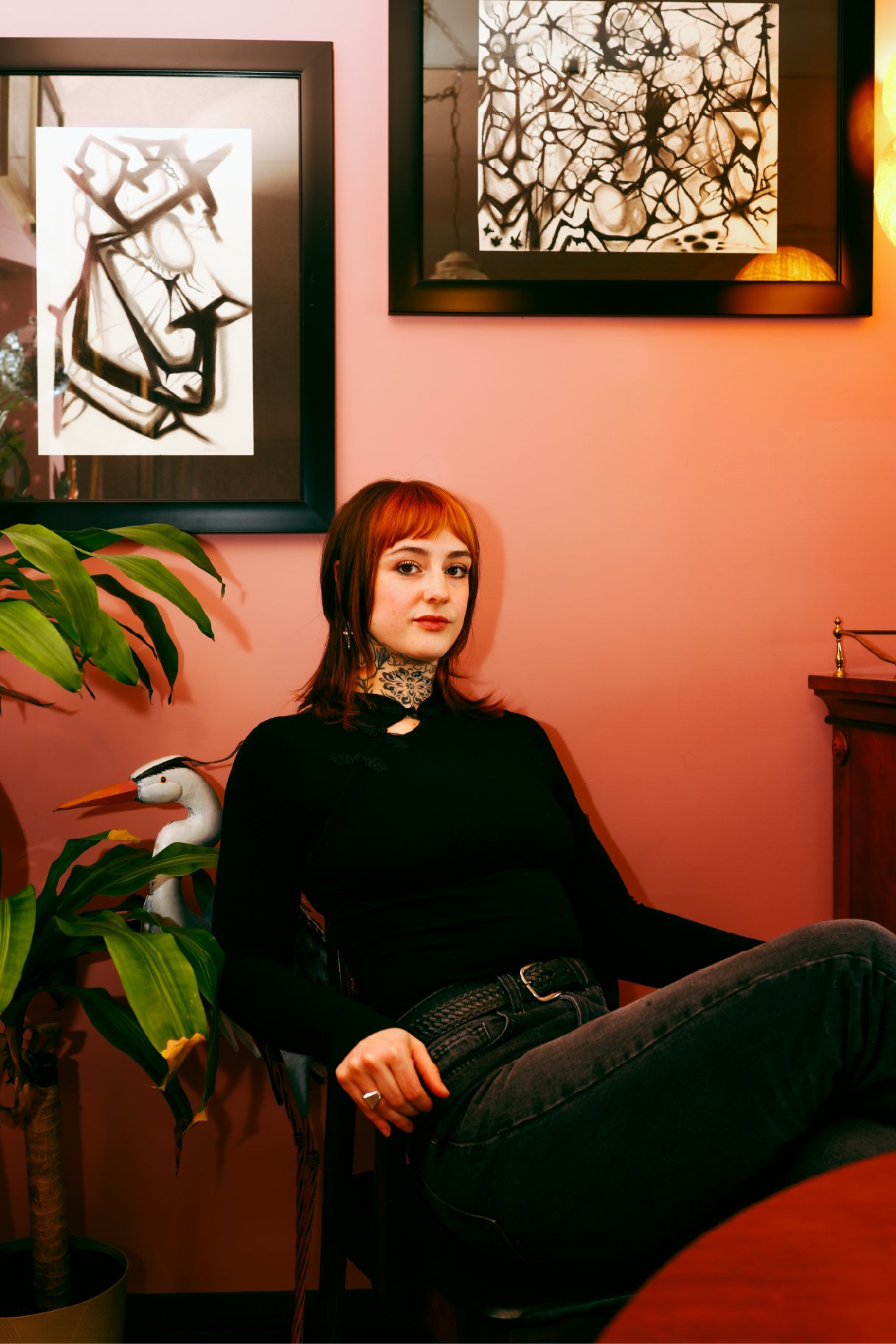 A young woman with red hair and tattoos on her neck, wearing a black top and black jeans, sitting on a chair in a warmly lit room with peach-colored walls. Behind her are two framed black and white abstract art pieces, a large plant to her left with 