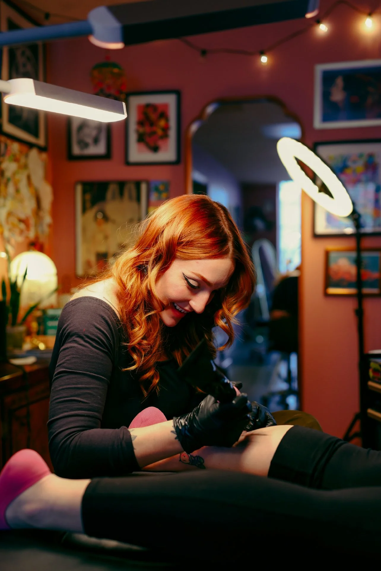 A woman with red hair and tattoos on her arm is tattooing a person's leg in a tattoo studio decorated with colorful artwork and warm lighting.