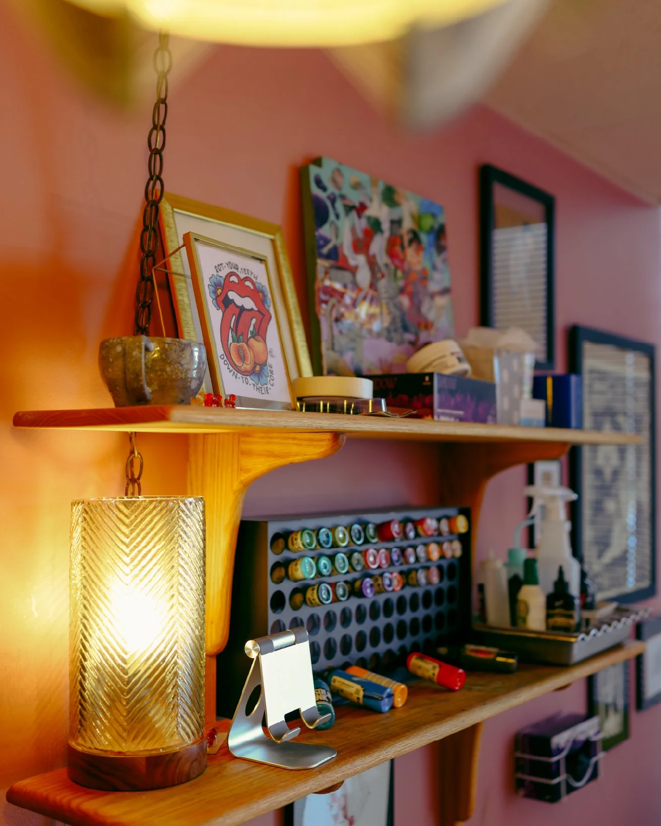 A wooden wall shelf with a textured glass lamp, framed artwork including The Rolling Stones logo, small art pieces, and art supplies such as paint bottles, brushes, and spray bottles. The background features a pink wall and a window with blinds.