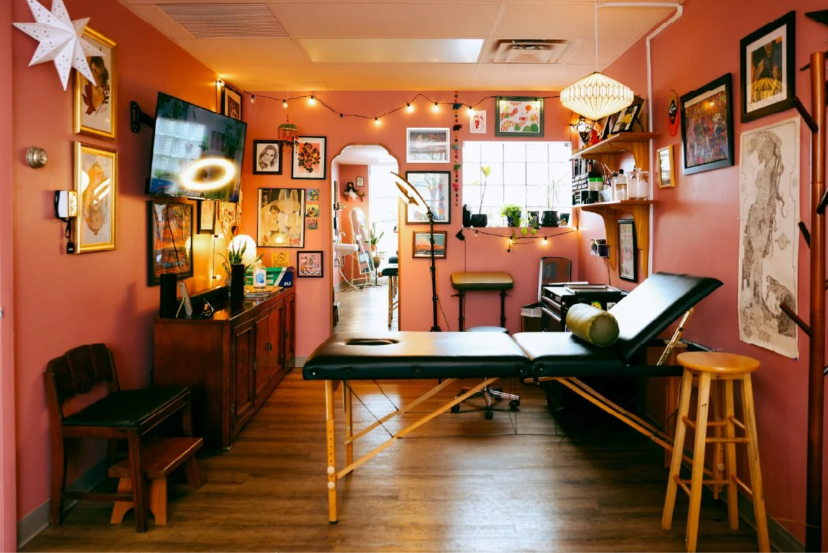 A cozy room set up for massage therapy with a massage table in the center, surrounded by various artwork, shelves, and potted plants, with soft lighting and a warm pink wall color.