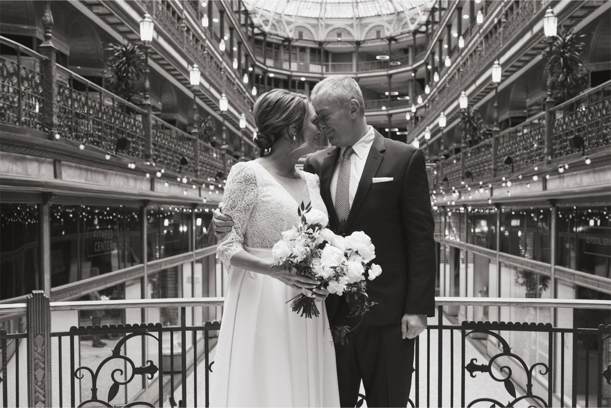 A black and white photo of a wedding couple standing close together in a multi-level mall, sharing a tender moment. The woman is holding a bouquet of flowers and wearing a lace wedding dress, while the man is in a suit and tie. They are smiling with