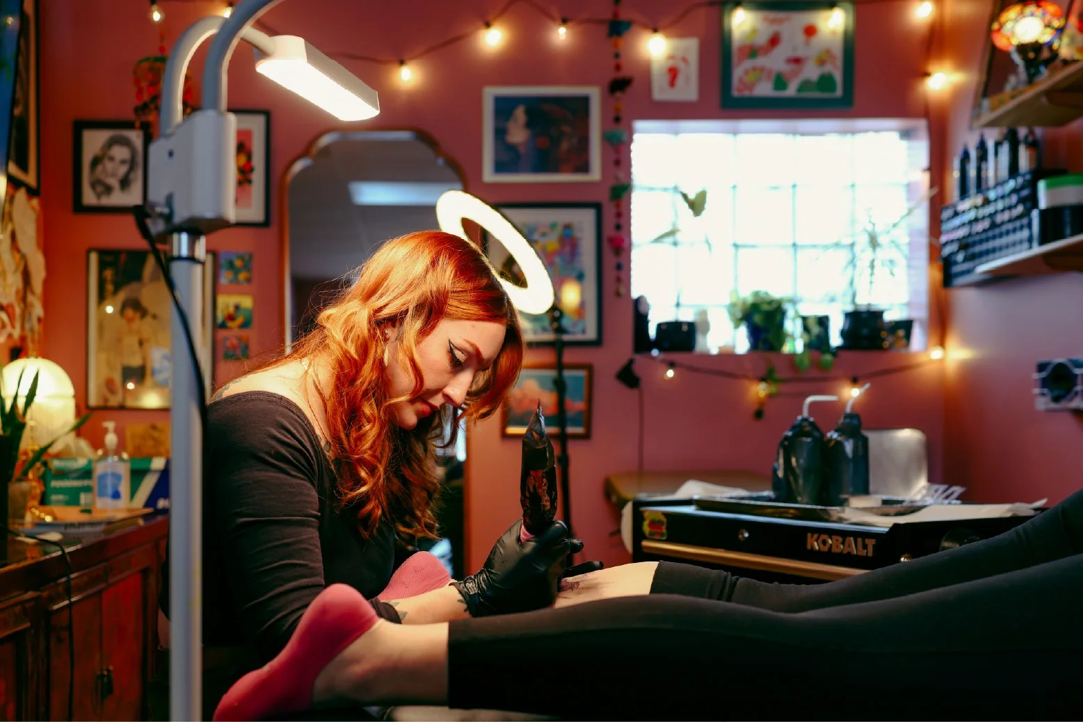 A woman with red hair tattoo, wearing black gloves, is tattooing a client in a colorful tattoo studio with pink walls, framed artwork, and string lights.