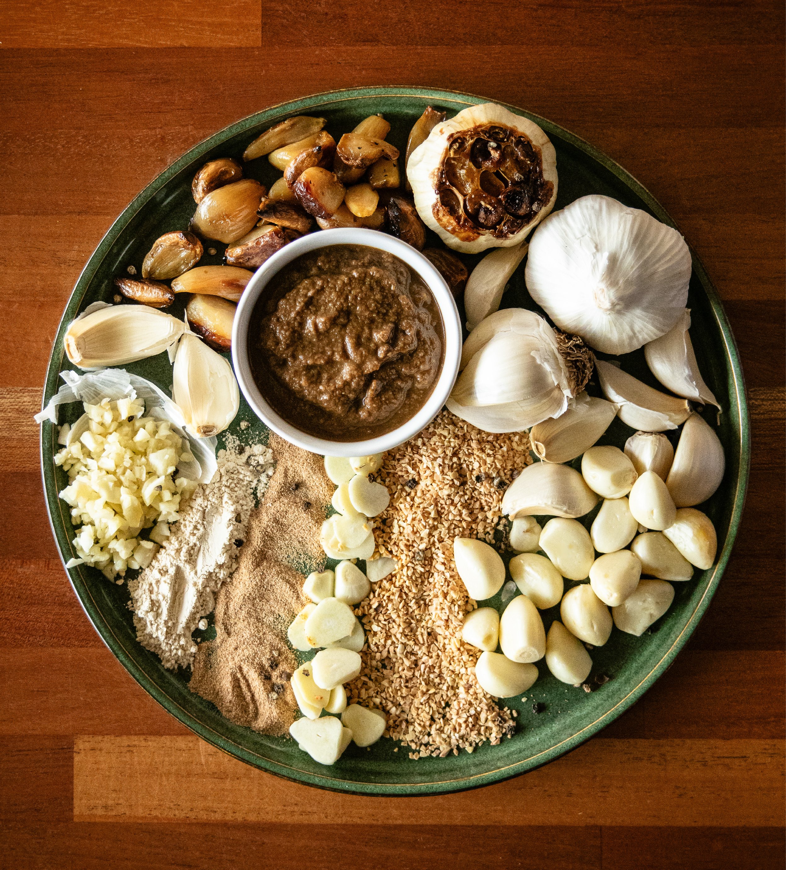 A large round tray with various ingredients for a garlic-themed dish. It includes garlic cloves, roasted garlic bulbs, chopped garlic, garlic powder, garlic salt, mashed garlic, a bowl of minced garlic paste, chopped nuts, and a jar of brown sauce or