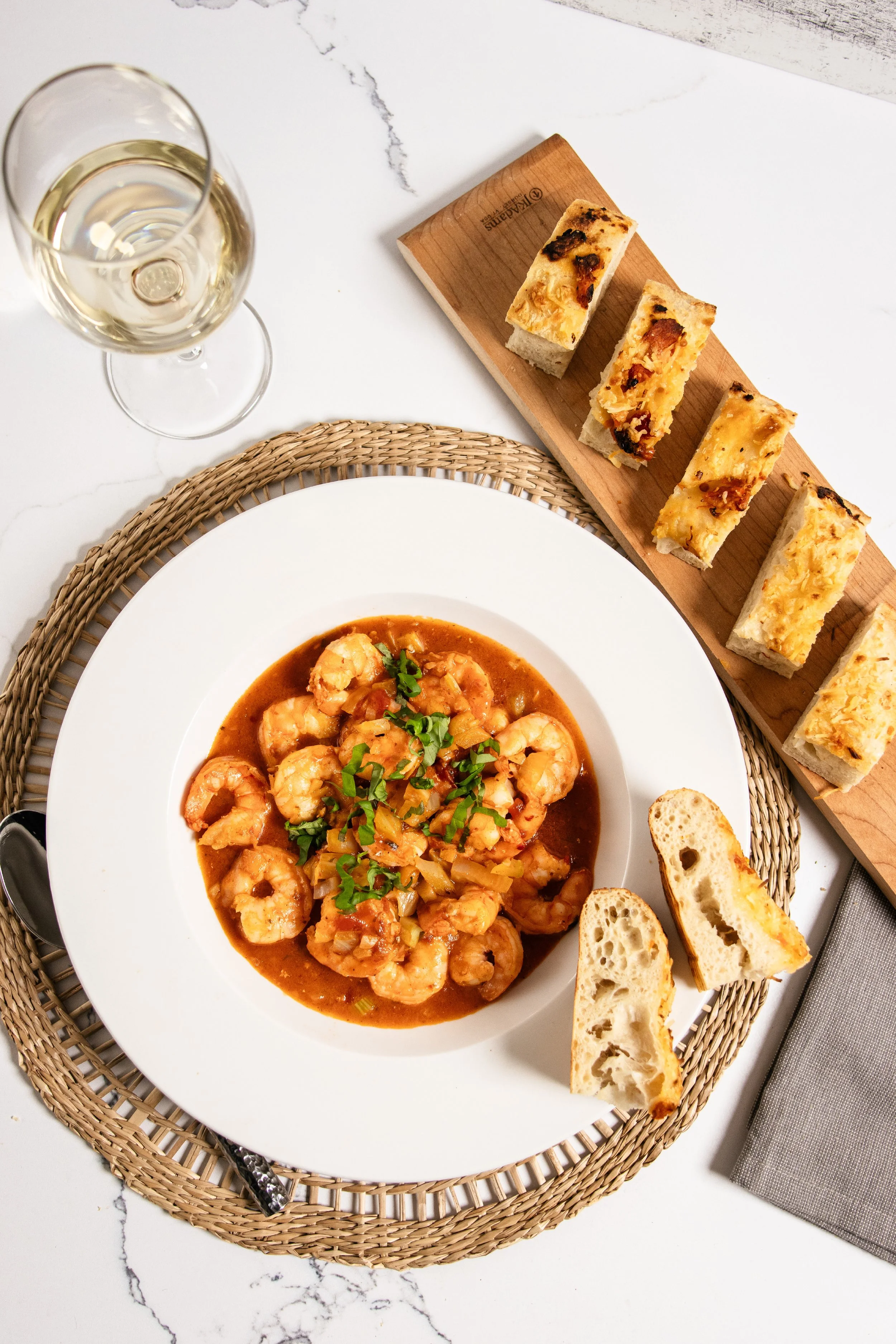 A plate of shrimp in sauce garnished with chopped herbs, accompanied by slices of toasted bread. A glass of white wine and a napkin are also visible on a white marble surface with a woven placemat.