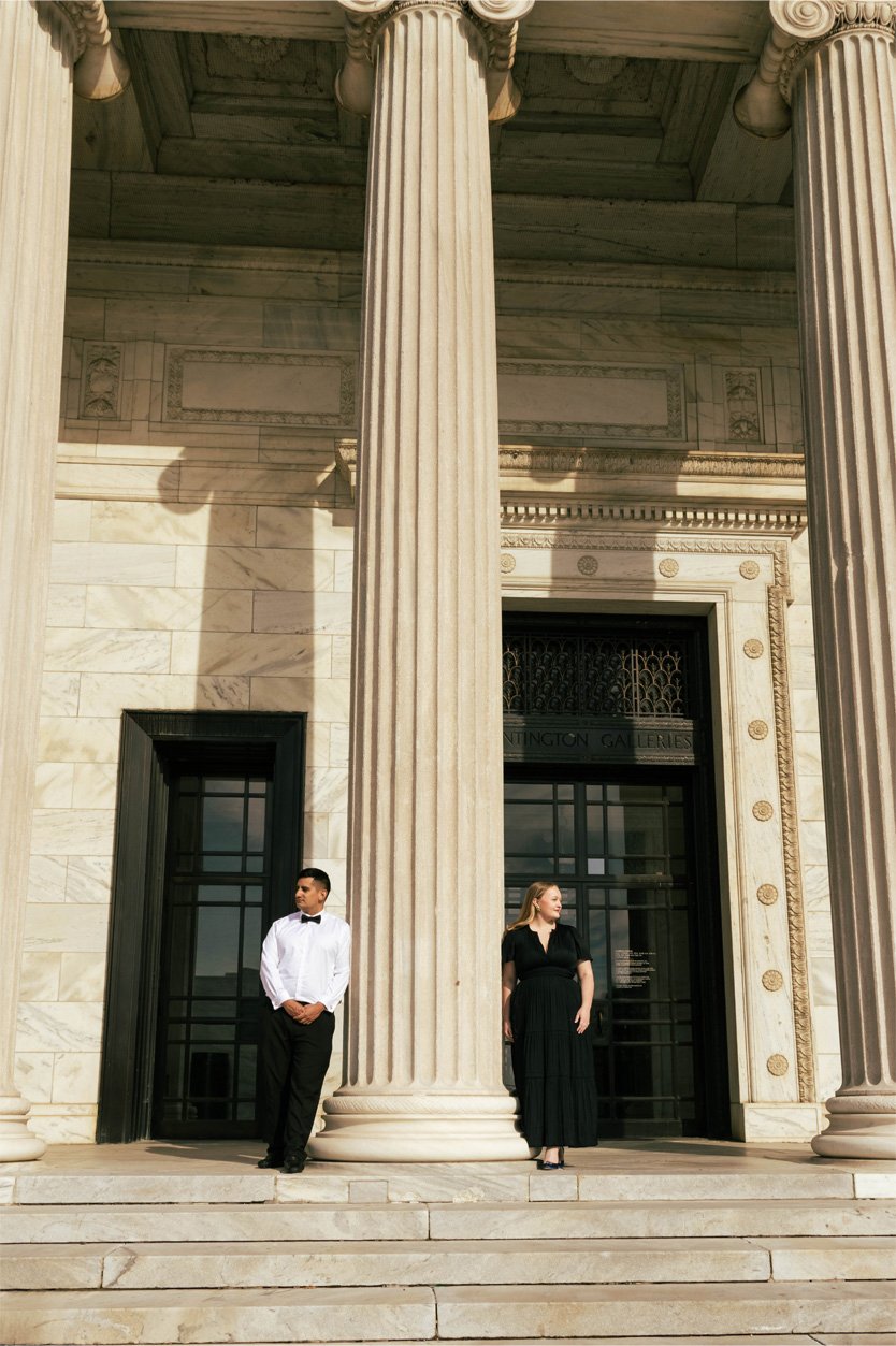 A man in a white shirt and black bow tie and a woman in a long black dress stand on steps in front of a neoclassical building with large columns and marble facade.