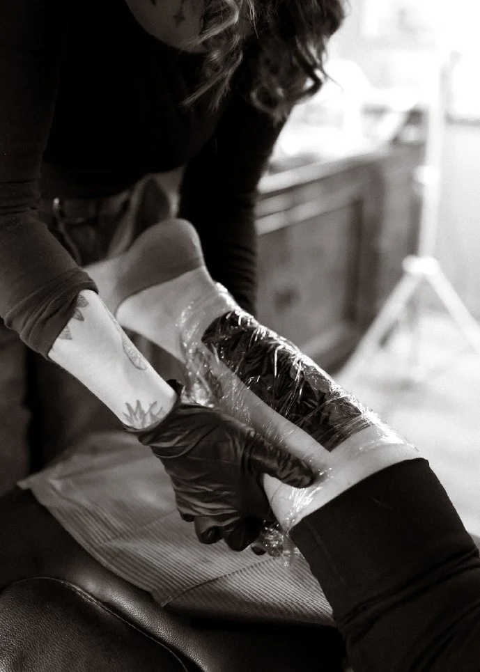 A person wearing black gloves is applying a tattoo or tattooing a person's arm, which is wrapped in plastic, in a studio or clinic with a wooden cabinet and blurred background.