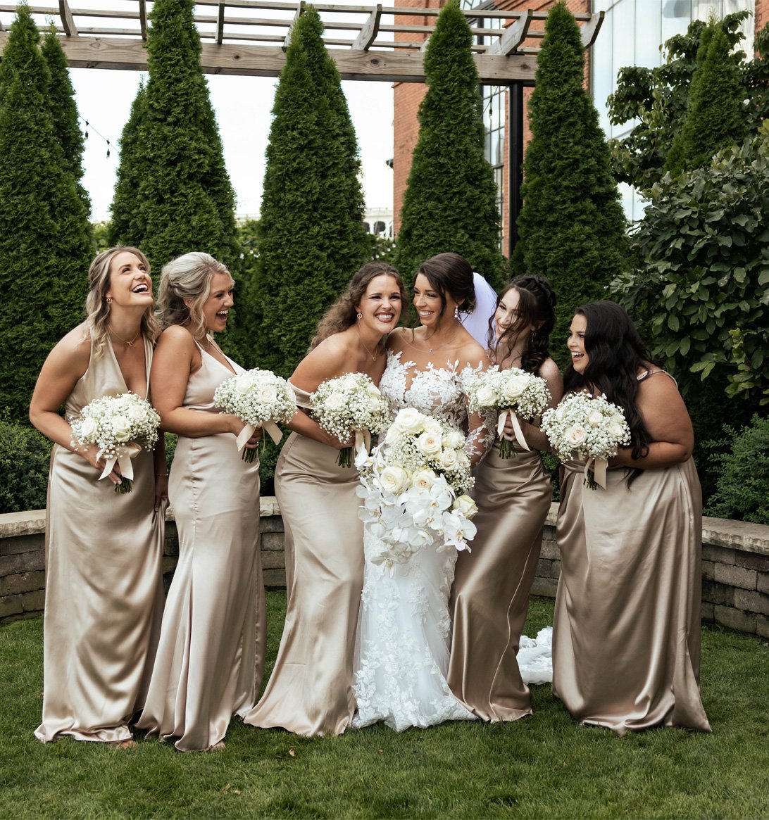 A bride and five bridesmaids in matching beige satin dresses, all smiling and laughing outdoors with green trees and a brick building in the background. The bride is holding a large bouquet of white flowers, and each bridesmaid is holding a smaller b