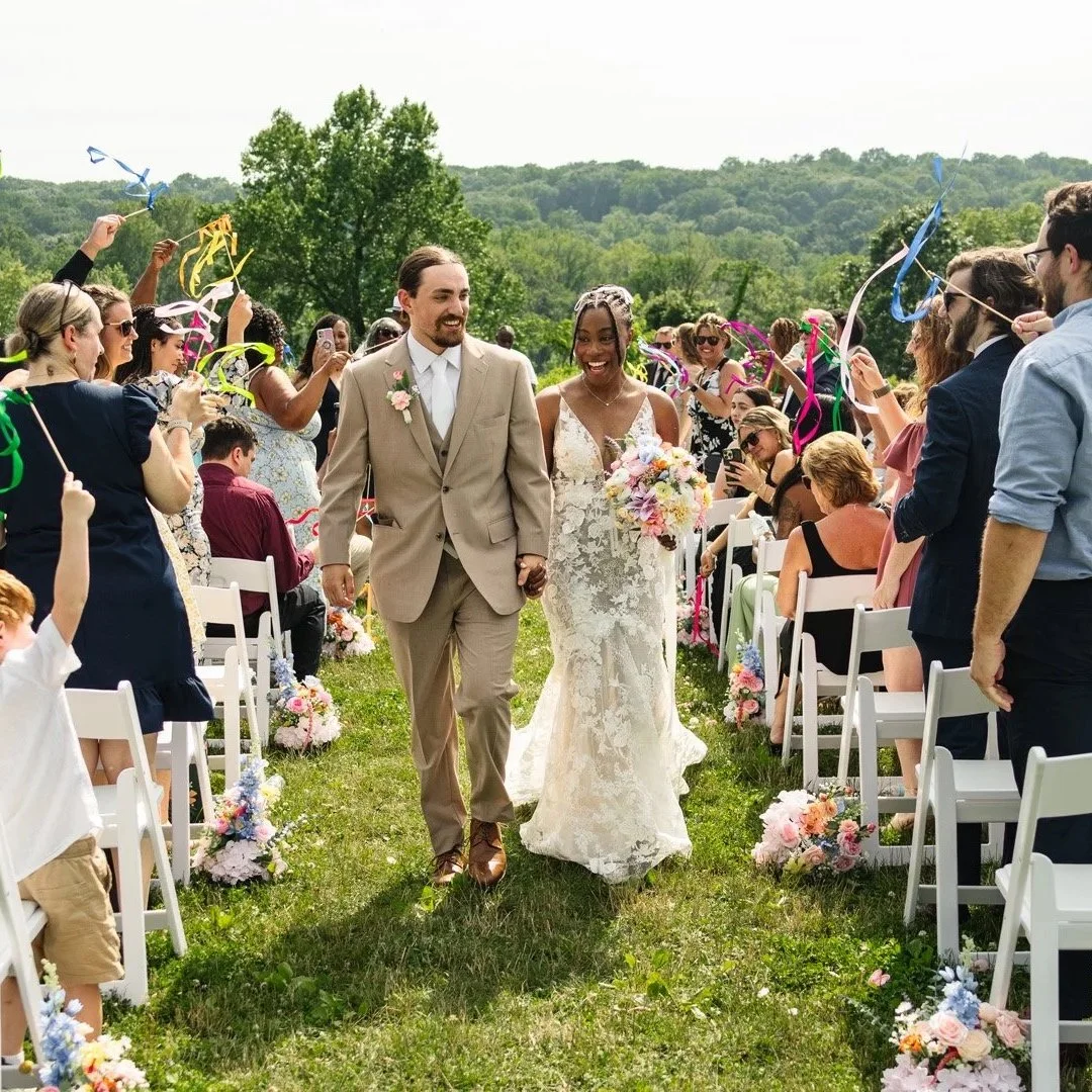 A newlywed couple walking down the aisle at an outdoor wedding, surrounded by smiling guests holding colorful streamers in a lush green setting.
