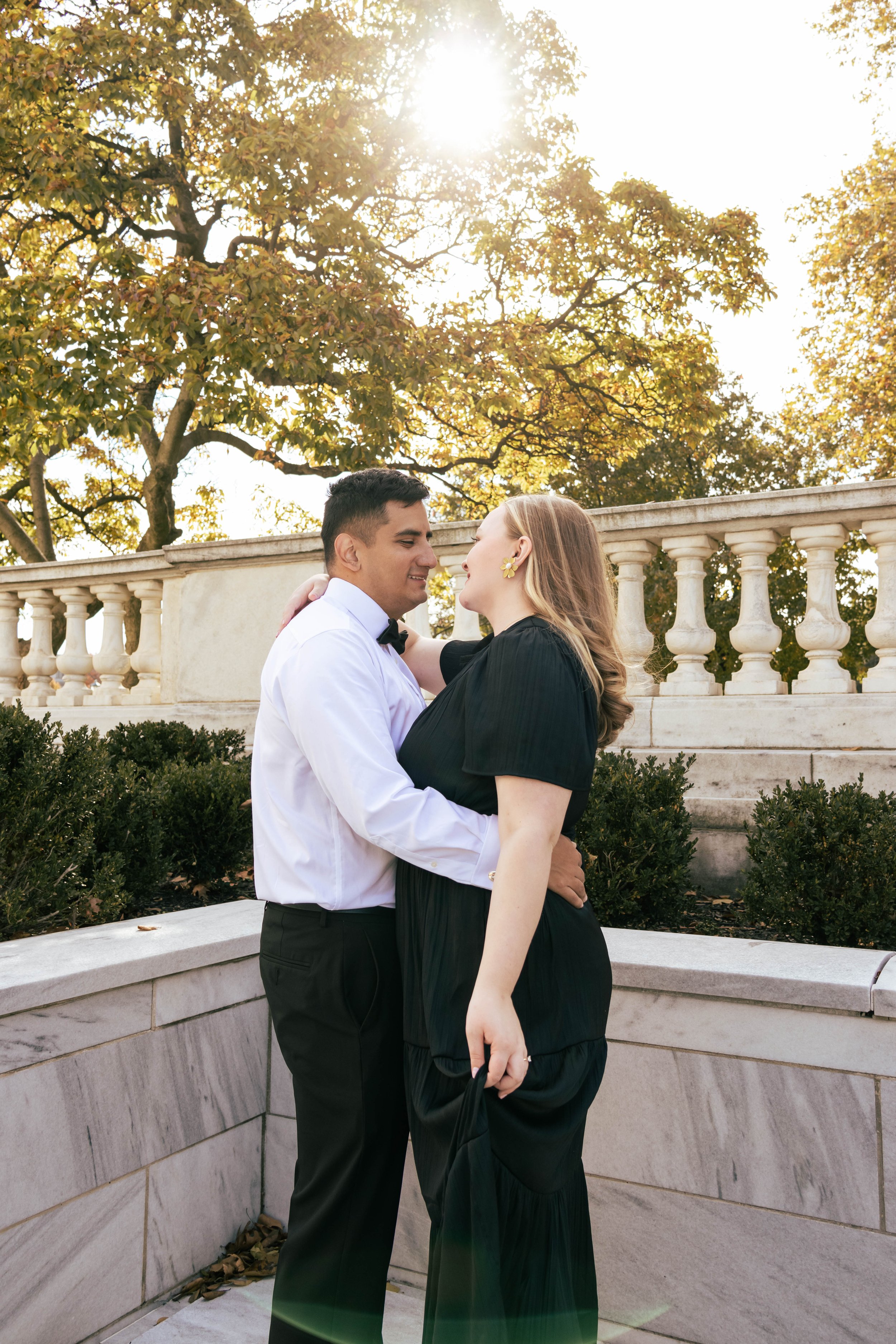 A couple in formal attire embracing outdoors at sunset, with trees and a stone railing in the background.