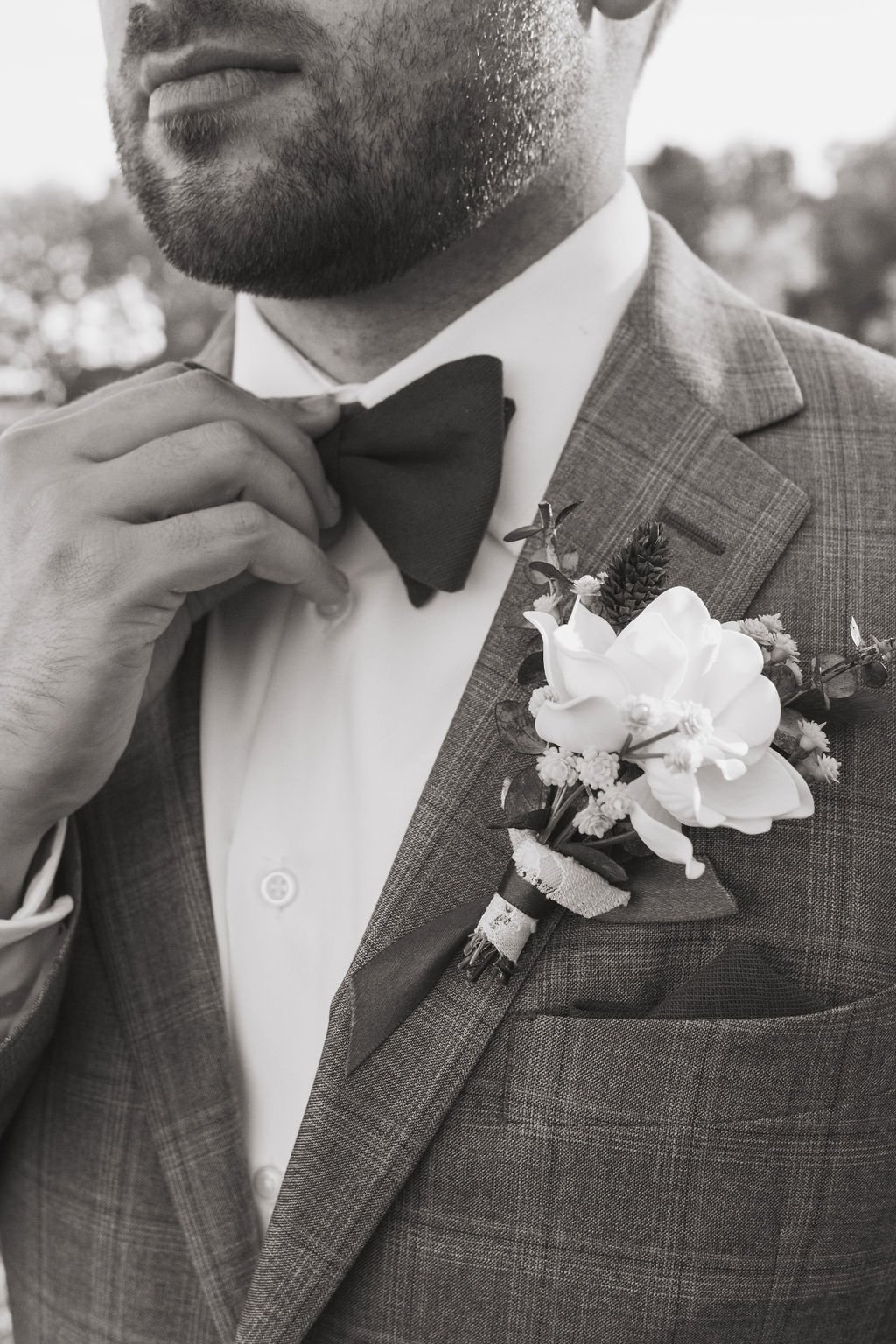 Close-up of a man adjusting a bow tie on his tuxedo, with a floral boutonniere on his lapel, in black and white.