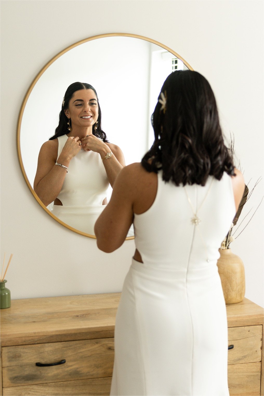 Woman in a white dress looking at herself in a round mirror, adjusting her necklace, with jewelry and a wooden table with decorated vases in a bright room.