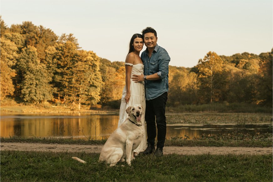 A couple standing on the grass by a lake with their yellow Labrador dog, surrounded by trees with autumn foliage, during sunset.