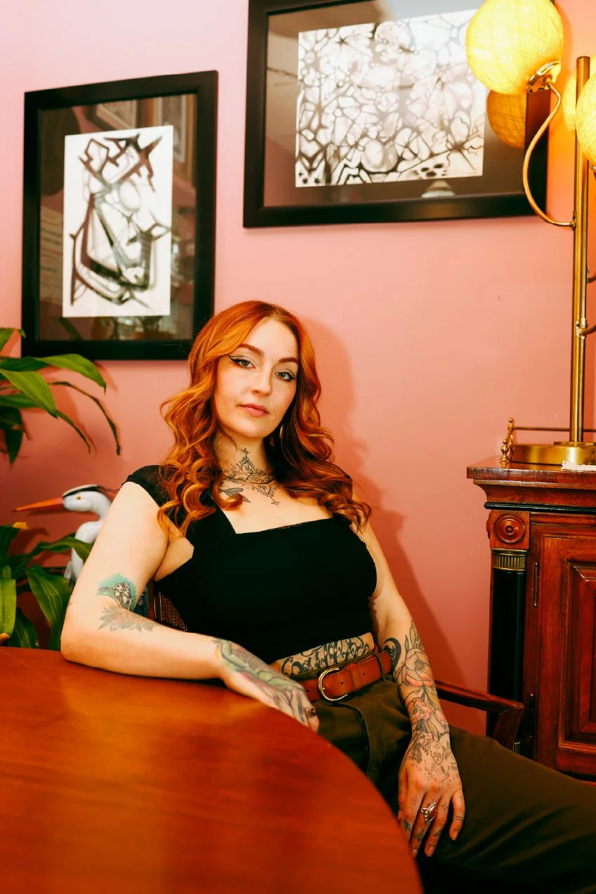 A young woman with red hair, tattoos on her arms and chest, sitting at a wooden table in a room with pink walls, black and white artwork on the wall, a wooden cabinet, green plant, and yellow lamps.