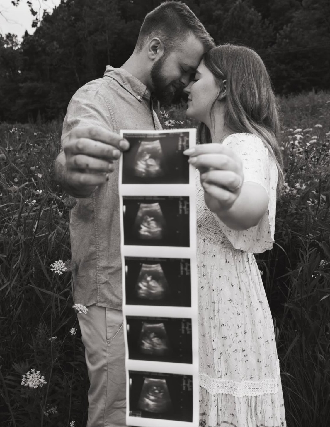 A couple holding ultrasound images in a field with wildflowers, their foreheads touching in a tender moment, both smiling softly.