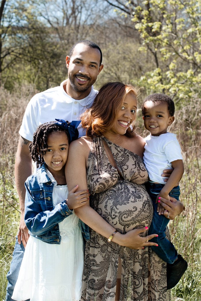 A happy family of four outdoors in a grassy field with trees, consisting of a pregnant woman, a man, a young girl, and a young boy, all smiling at the camera.