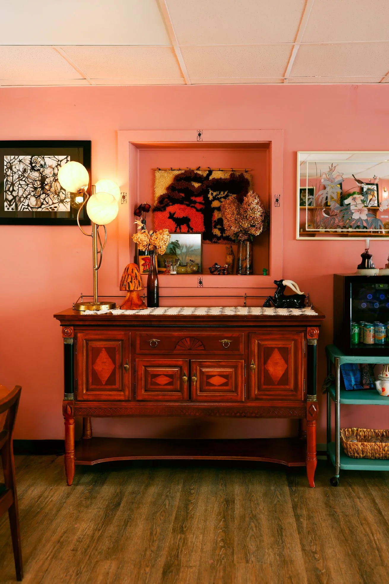 Vintage wooden sideboard with decorative inlay, topped with a floral lace runner, lamp, vase with flowers, and a small sculpture, against pink walls with artwork and mirror, on a hardwood floor.