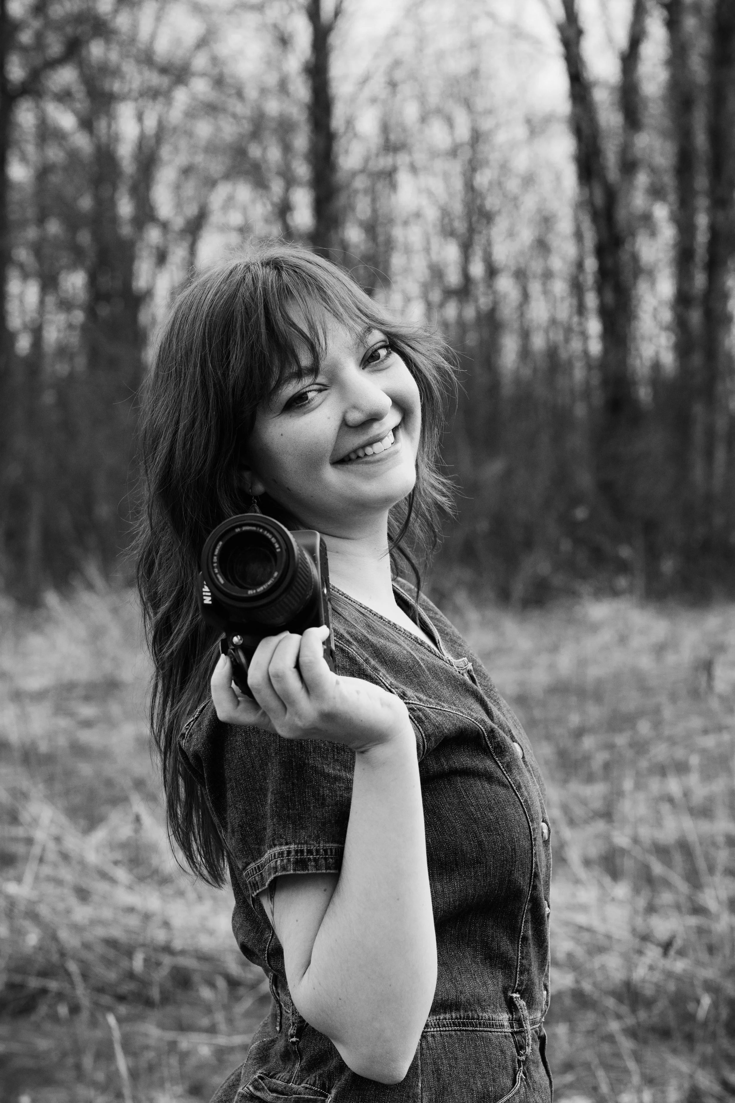 A young woman smiling, holding a camera, standing outdoors in a wooded area, black and white photograph.