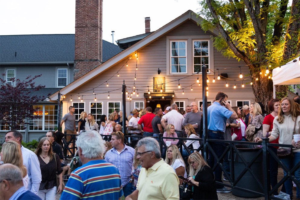 An outdoor gathering with many people socializing under string lights in front of a house.